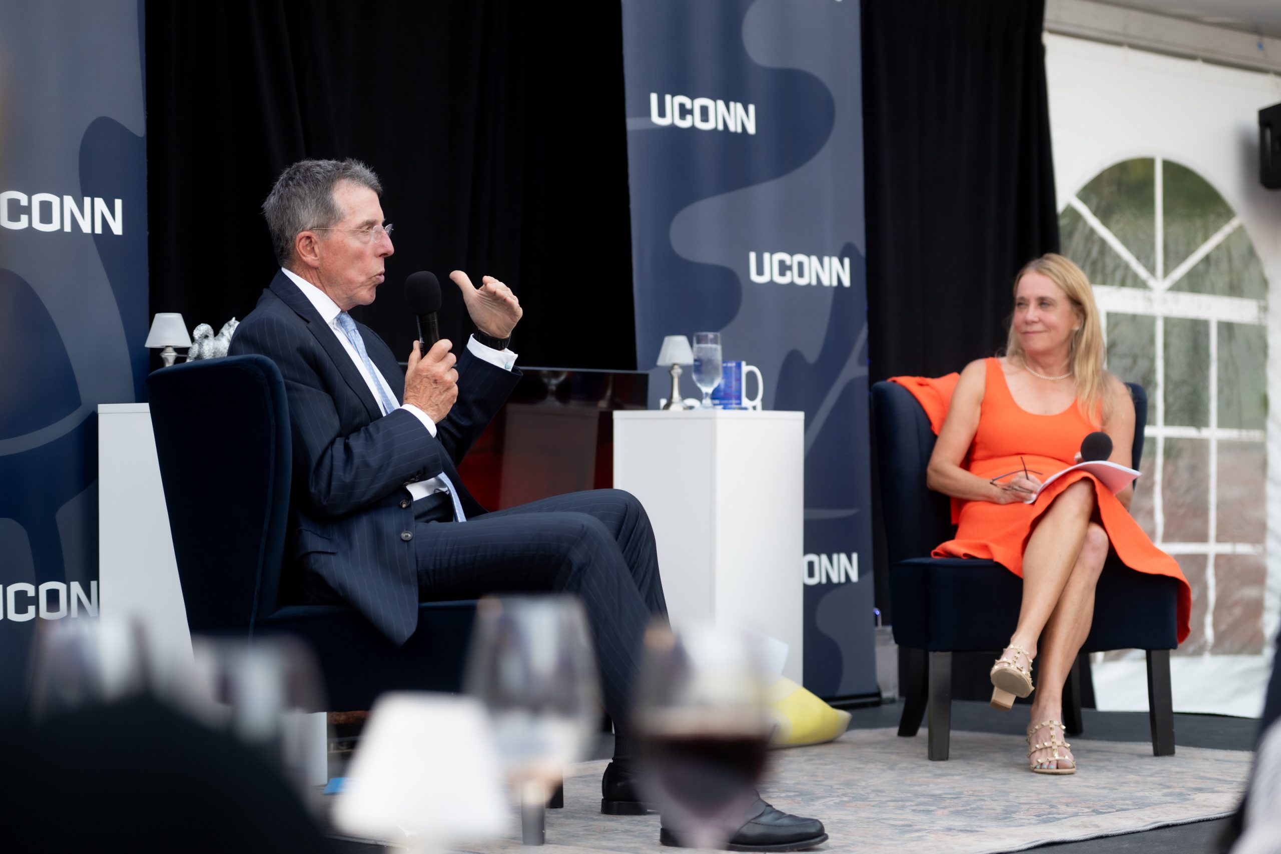 A man in a suit and a woman in an orange dress sit on stage with a small table between them and a UConn-branded backgrop behind them.