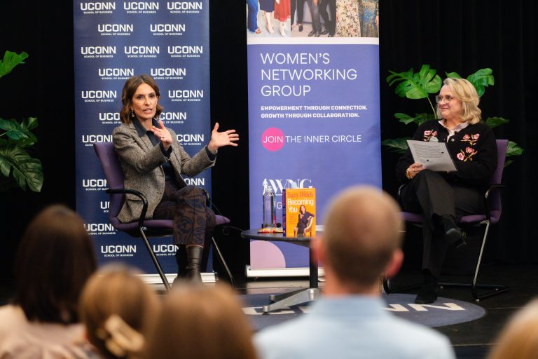 A woman in a gray blazer speaks to a crowd while seated on stage next to a woman in black. Behind them is a backdrop with UConn logos.