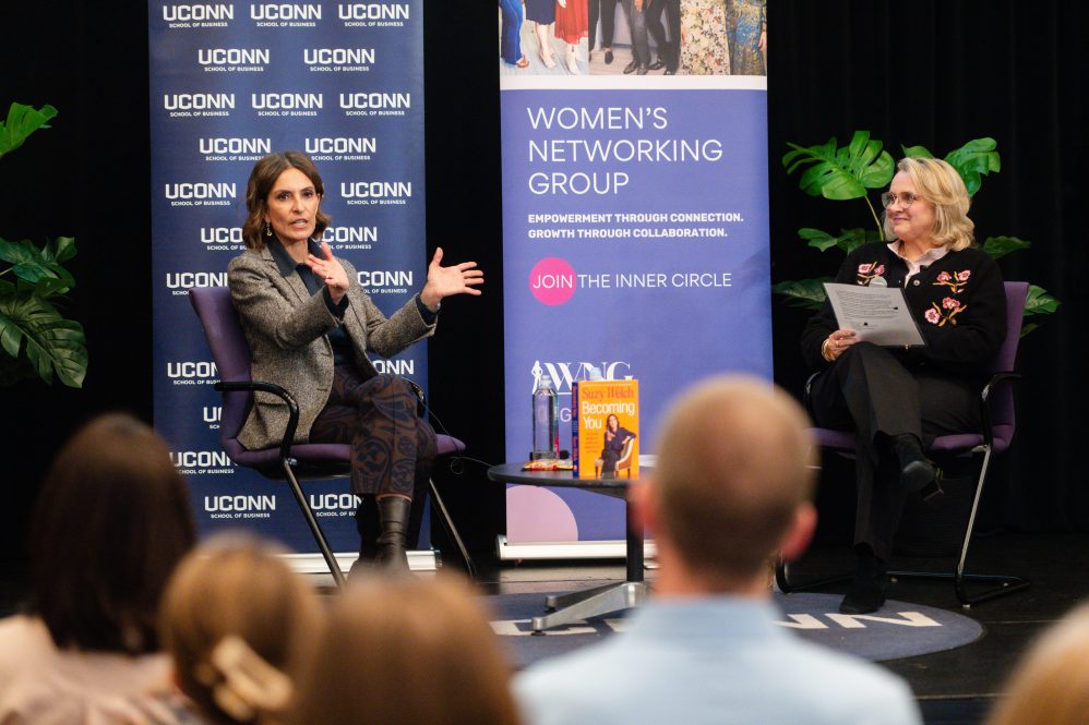 A woman in a gray blazer speaks to a crowd while seated on stage next to a woman in black. Behind them is a backdrop with UConn logos.