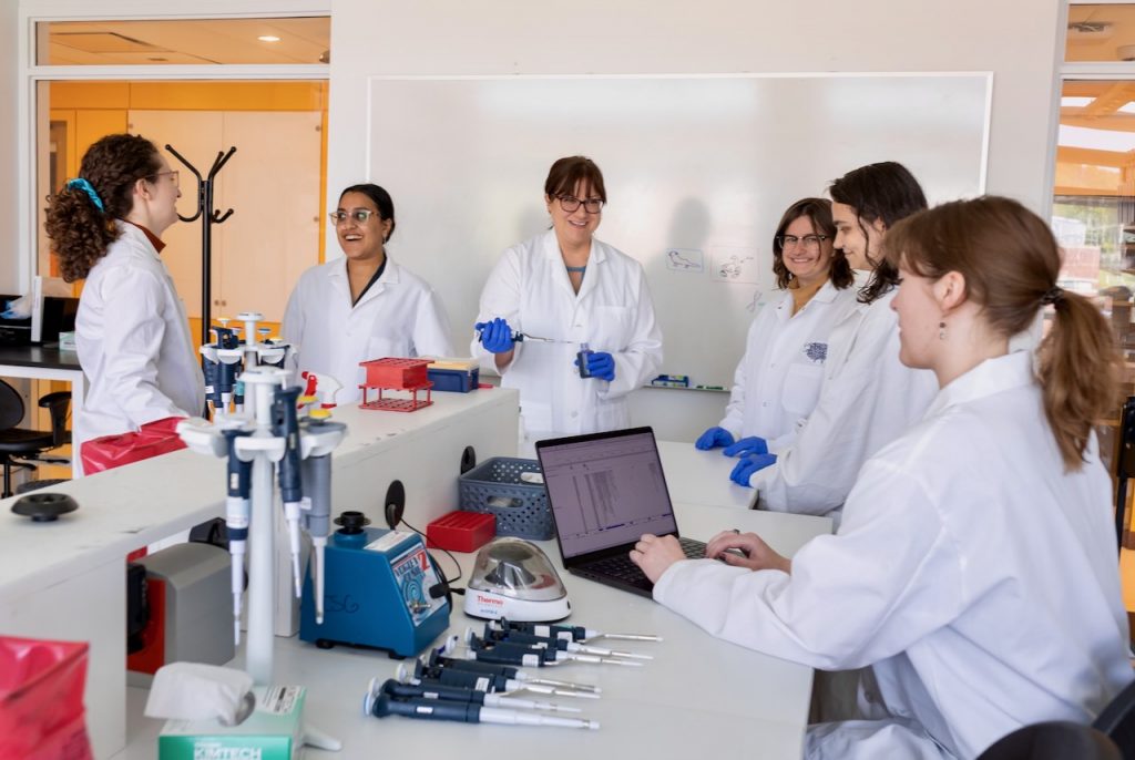 Six people in lab coats smile around a lab station
