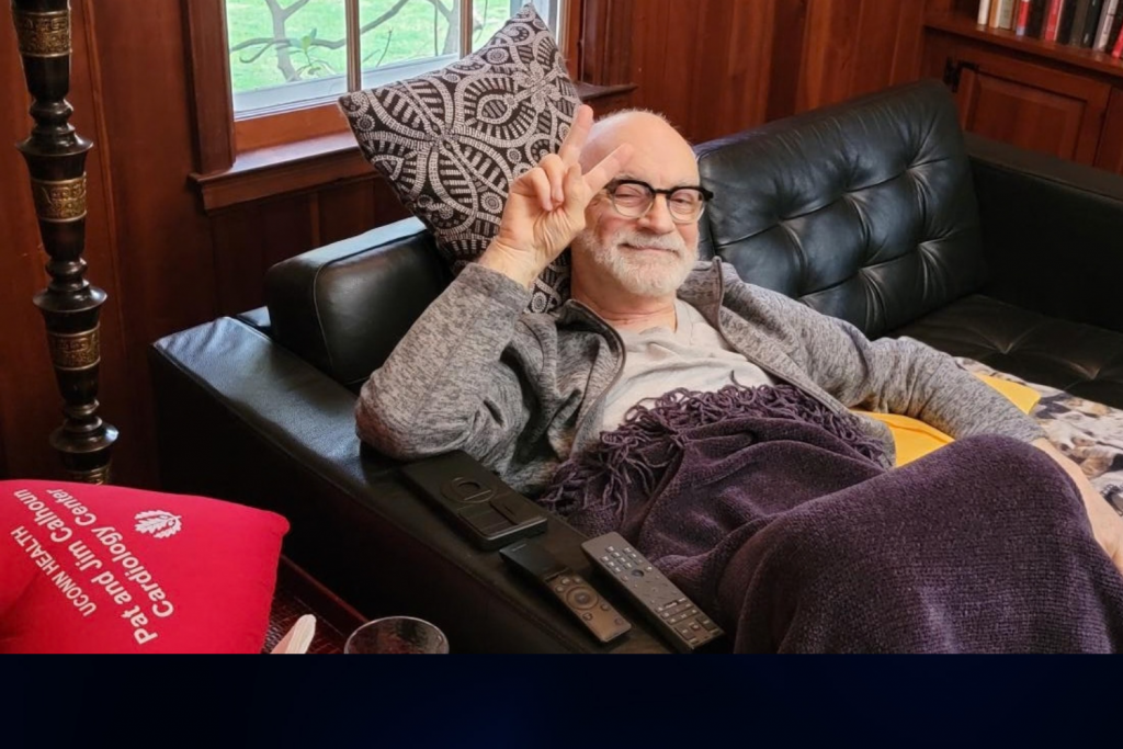 man laying on couch with blanket over him giving the peace sign with this hand