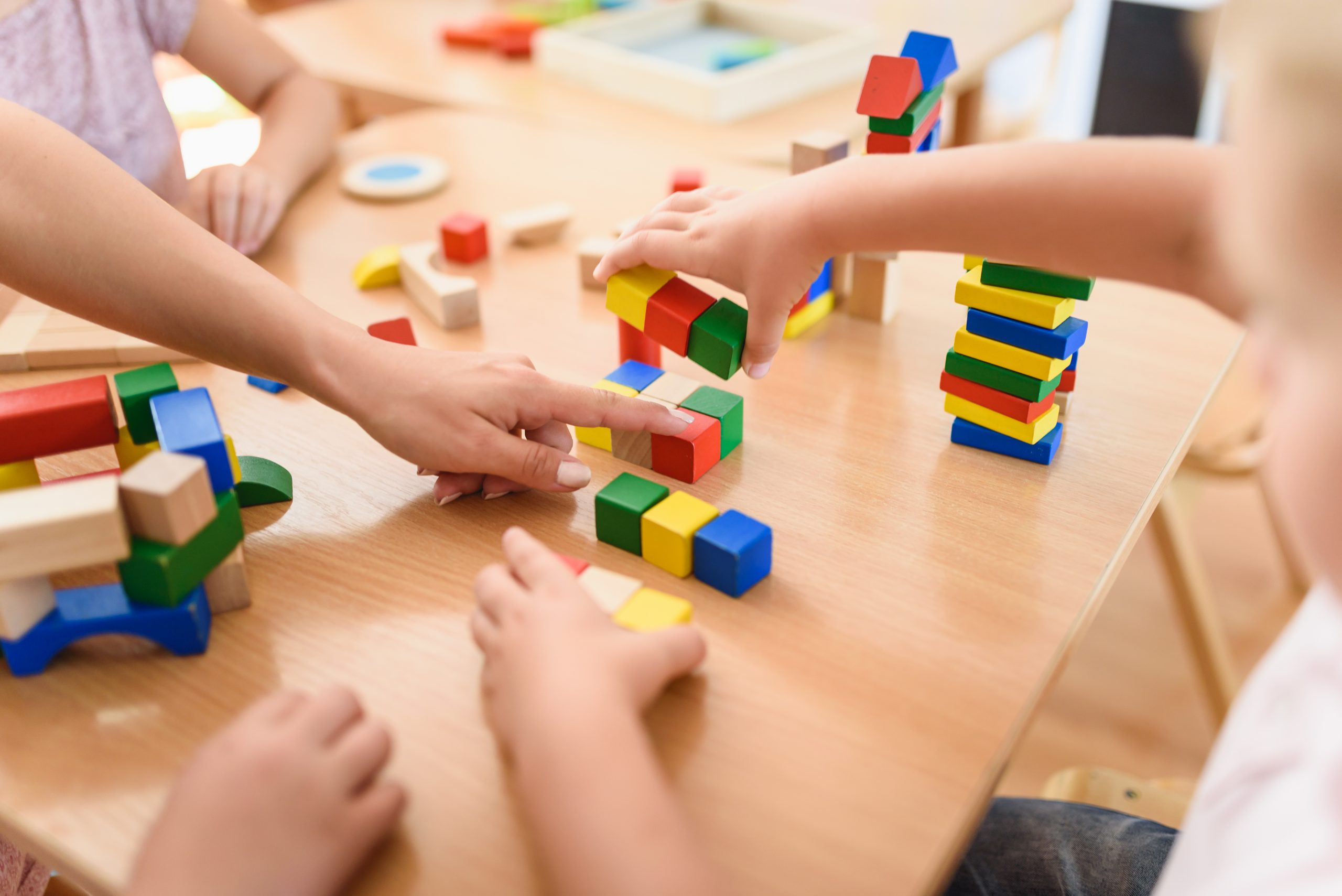 Young children's hands play with colorful blocks on a wooden table.