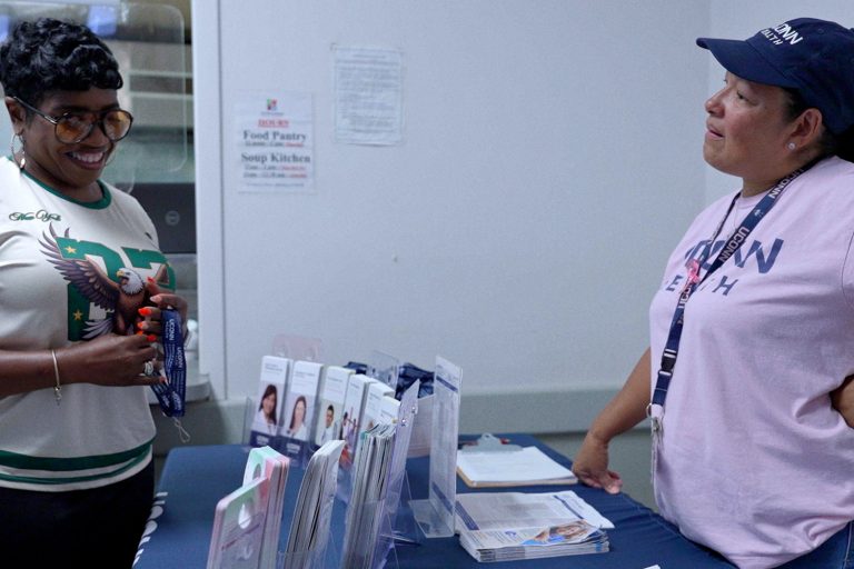 A woman in a baseball cap standing behind a table talking to another woman on the other side of the table with brochures on the table
