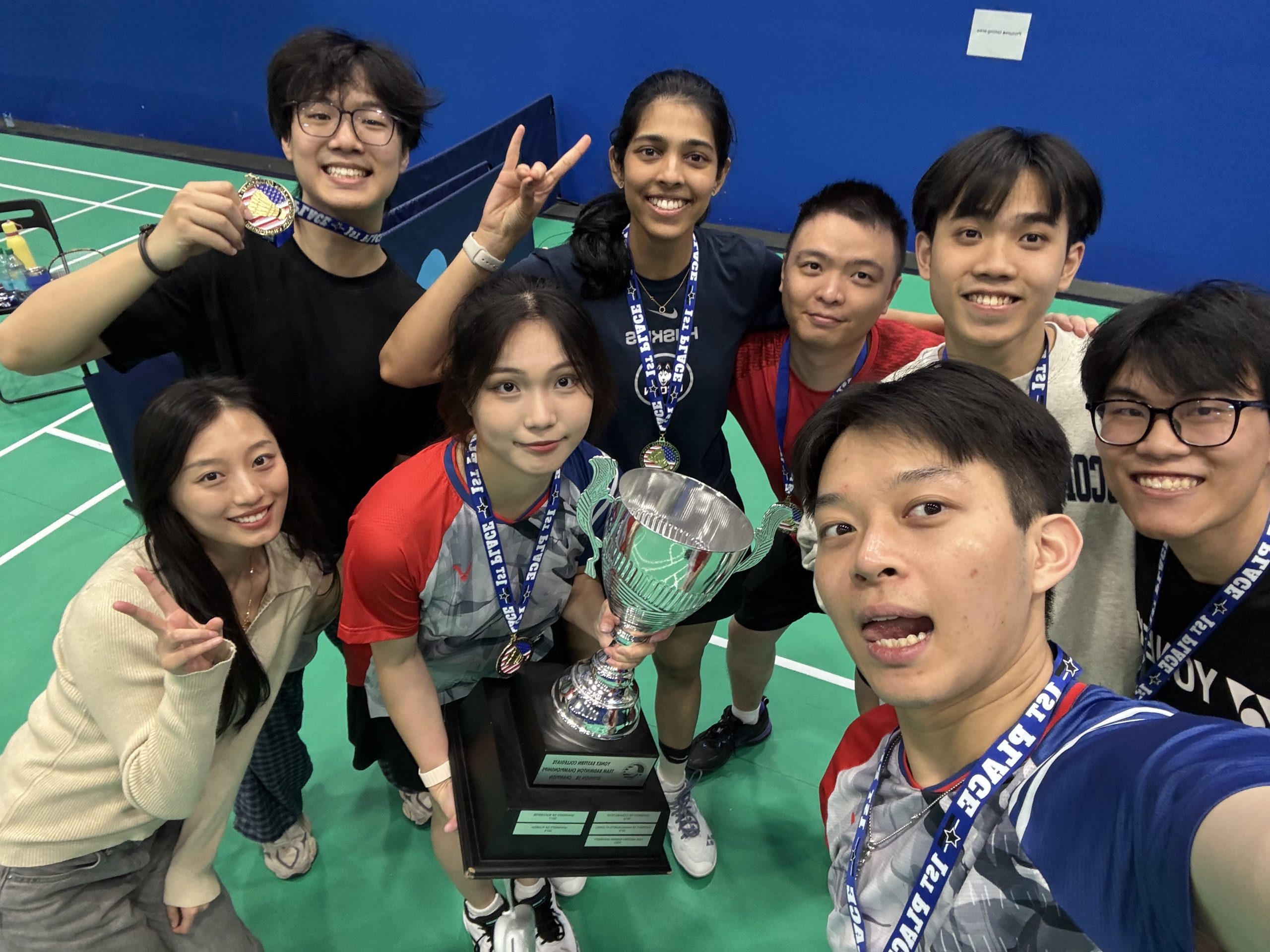 A group of students pose with a championship trophy on a badminton court.