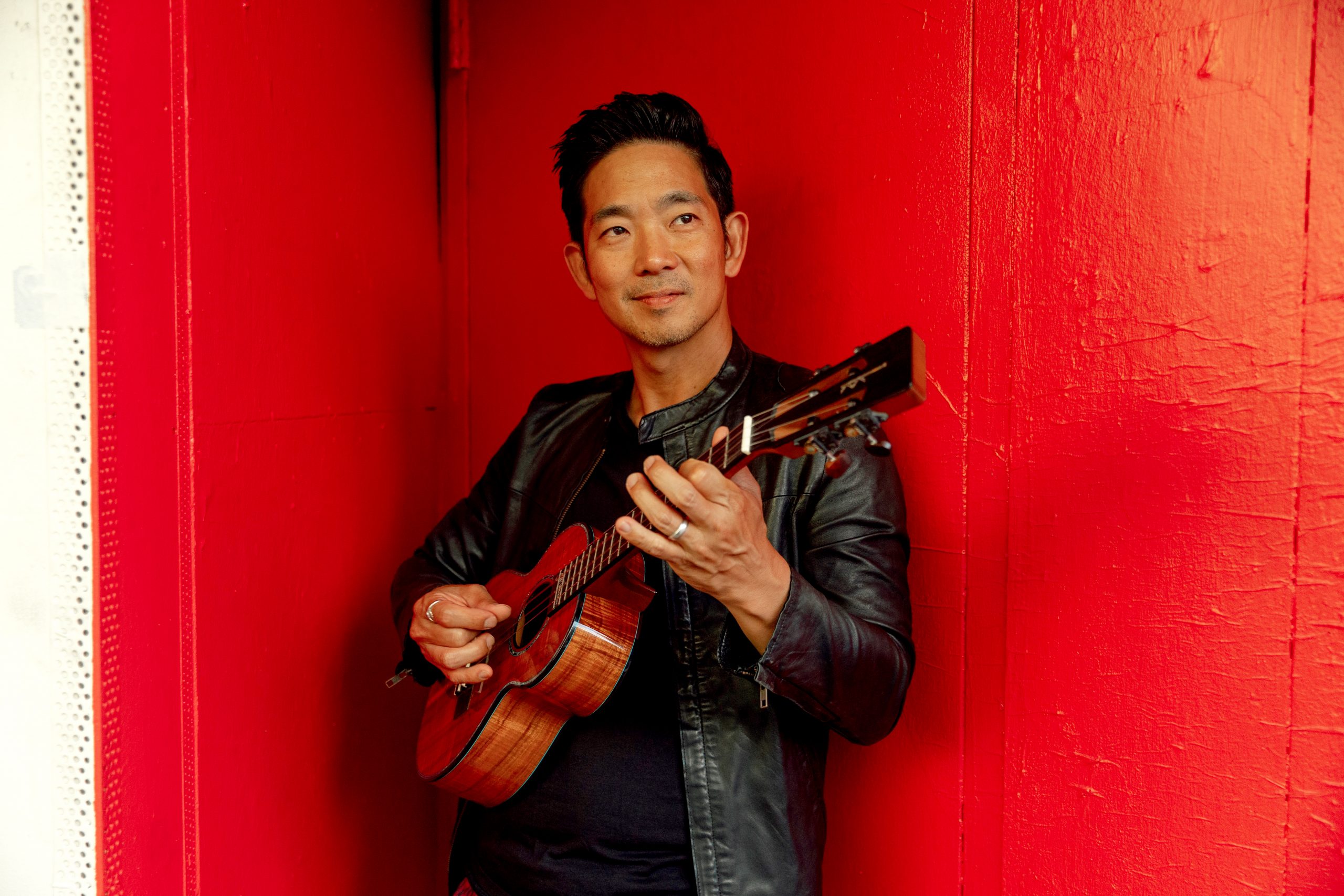 A man in a black jacket holds a ukulele in front of a red wall.