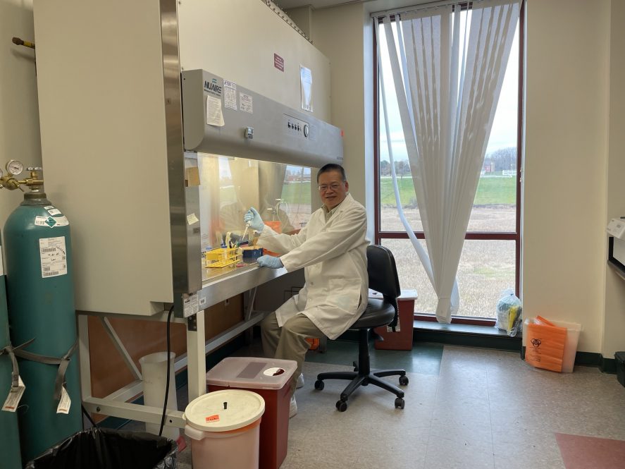 A man with glasses in a white lab coat and blue plastic gloves holds a test tube while sitting in a laboratory.