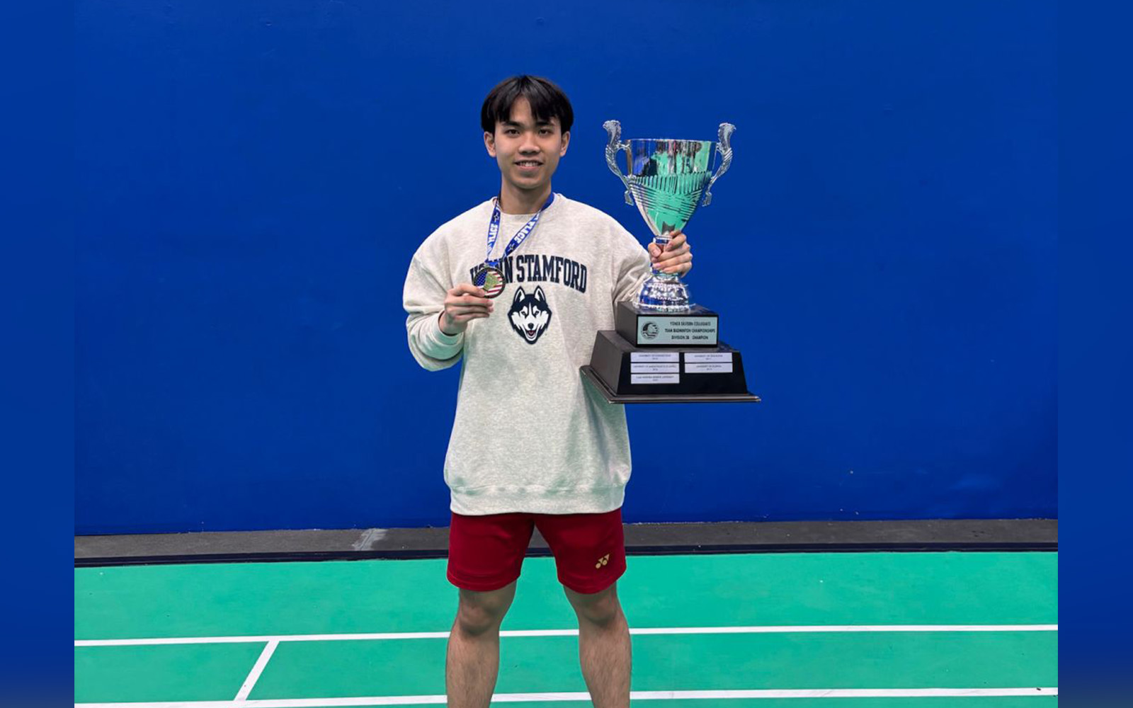 A student stands on a badminton court holding a championship trophy.
